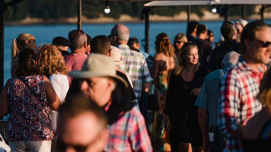 Full shot of a large crowd of people, likely at an outdoor event or gathering by a body of water.   The people are densely packed together, with various ages and styles of clothing.   Many people are wearing casual attire, including shirts, tops, and dresses, and some are visible with hats and sunglasses. 