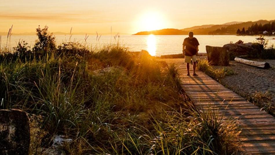 A man walks away from a waterfront along a wooden boardwalk as the sun sets behind him.